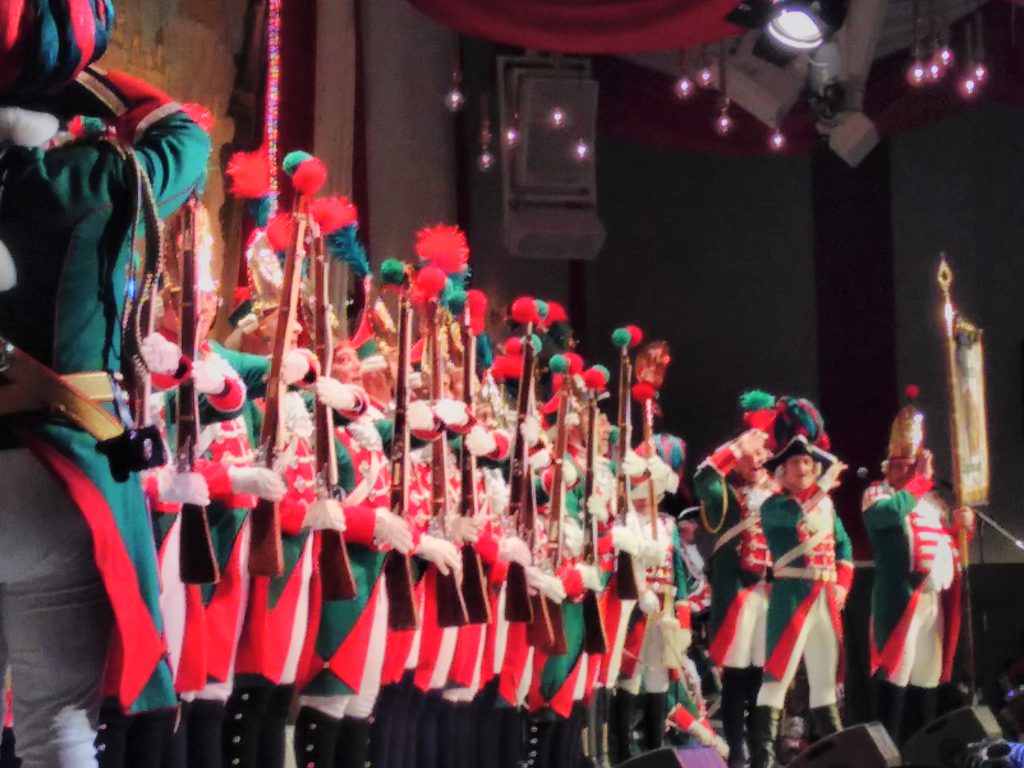 A line of men in 18th century soldier uniforms on stage at a Karneval Sitzung